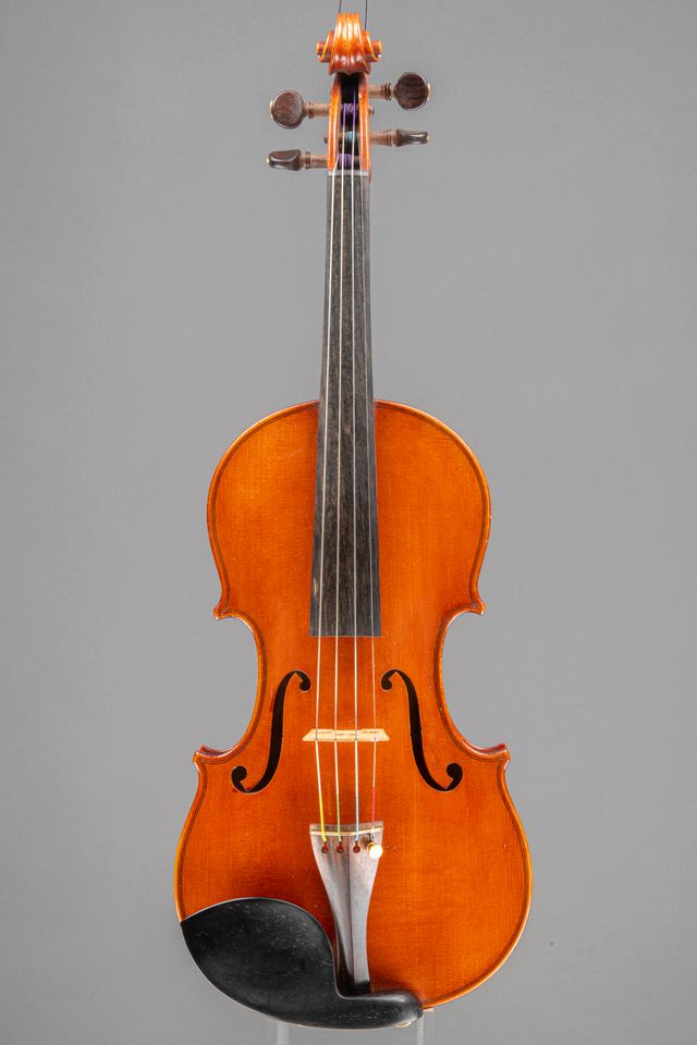 A full-length, front-facing view of a polished wooden violin shown against a neutral grey background.