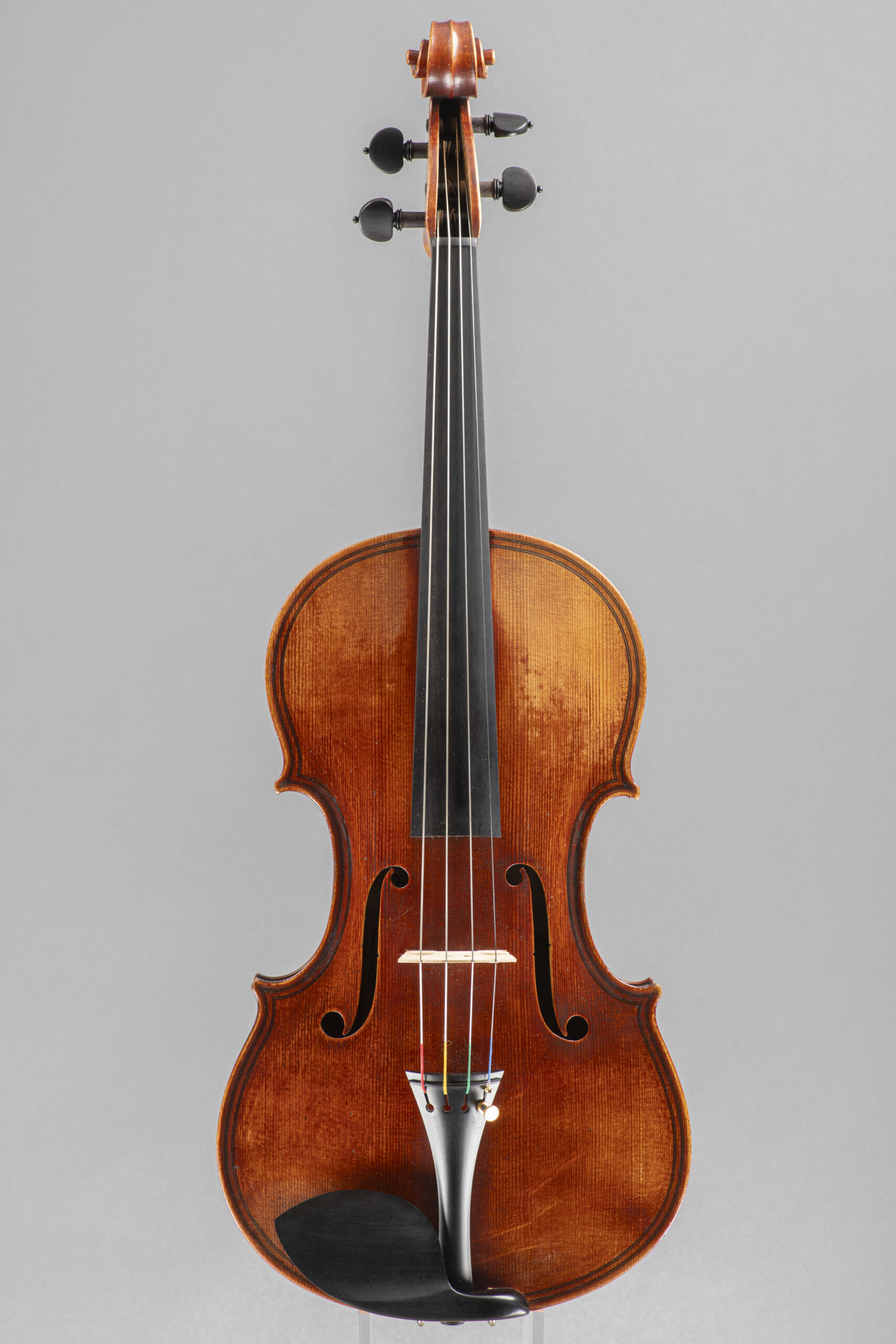 Front view of a vintage wooden violin, showcasing its deep amber varnish and black chin rest, positioned vertically on a grey backdrop.