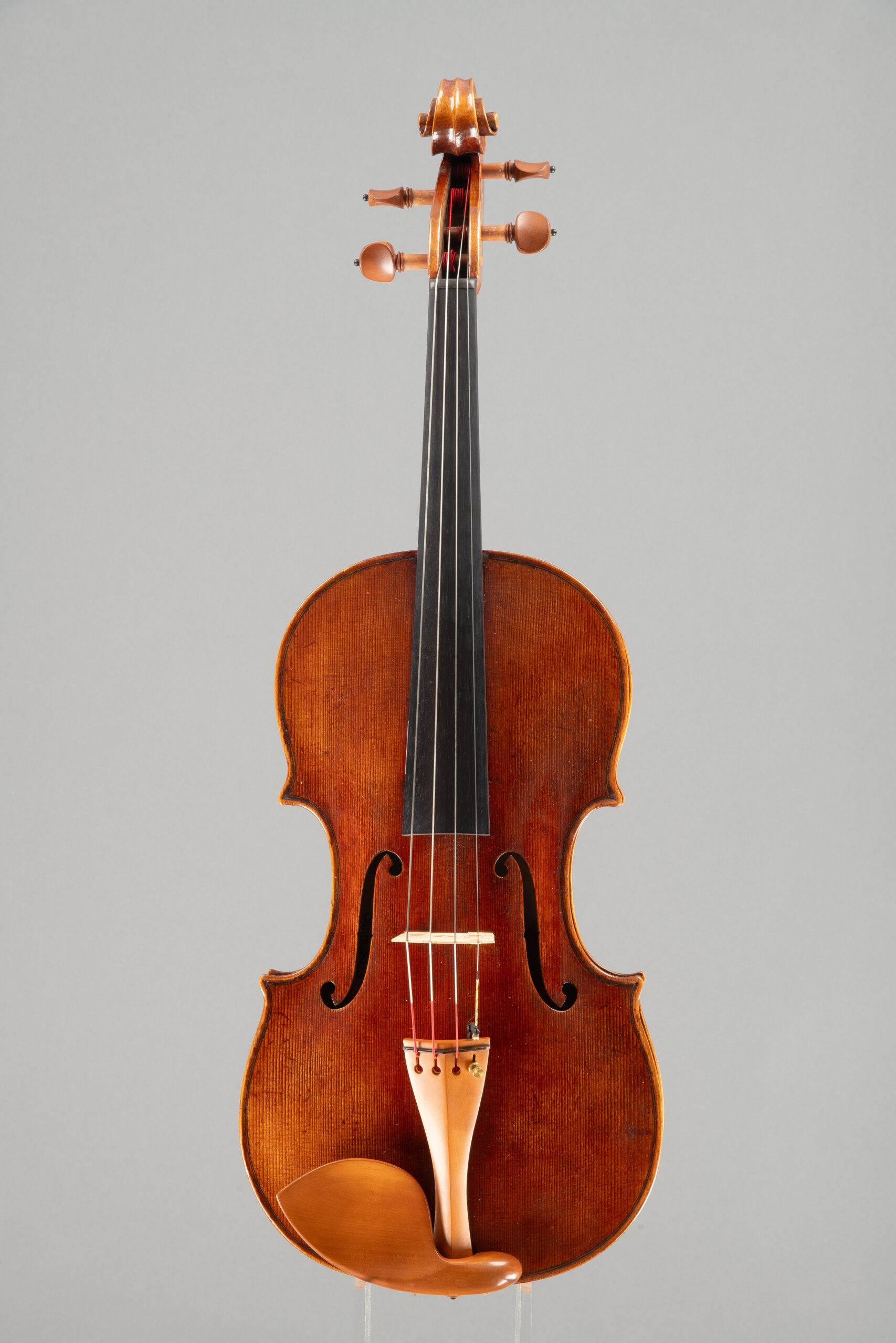 An upright, studio-lit photograph of a wooden violin featuring an aged, dark-toned varnish, a black fingerboard, and a lighter-colored chin rest, set against a plain grey backdrop.