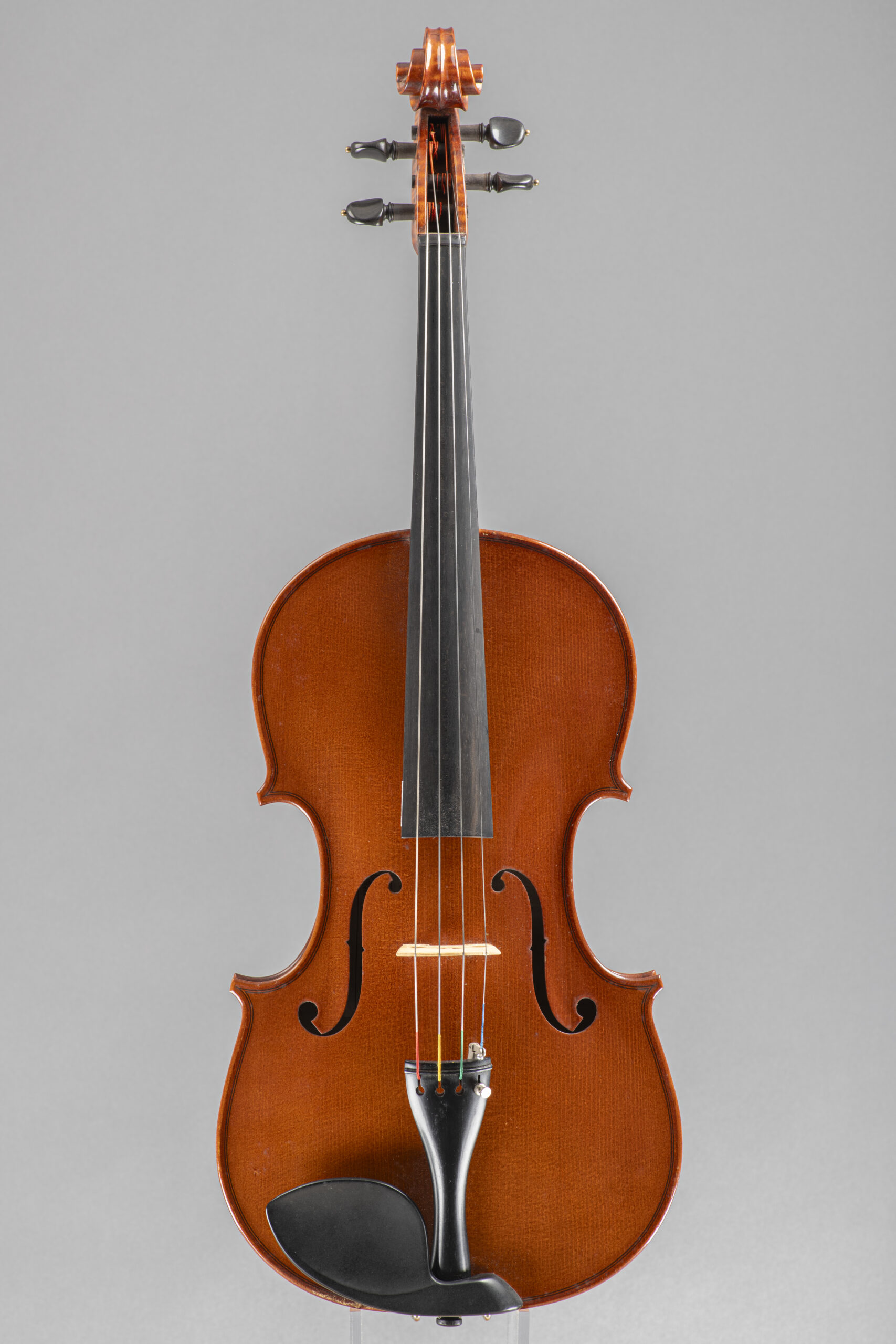 An upright studio portrait of a wooden violin featuring a classic polished finish, a dark ebony fingerboard, and a black chin rest, centered against a neutral grey backdrop.