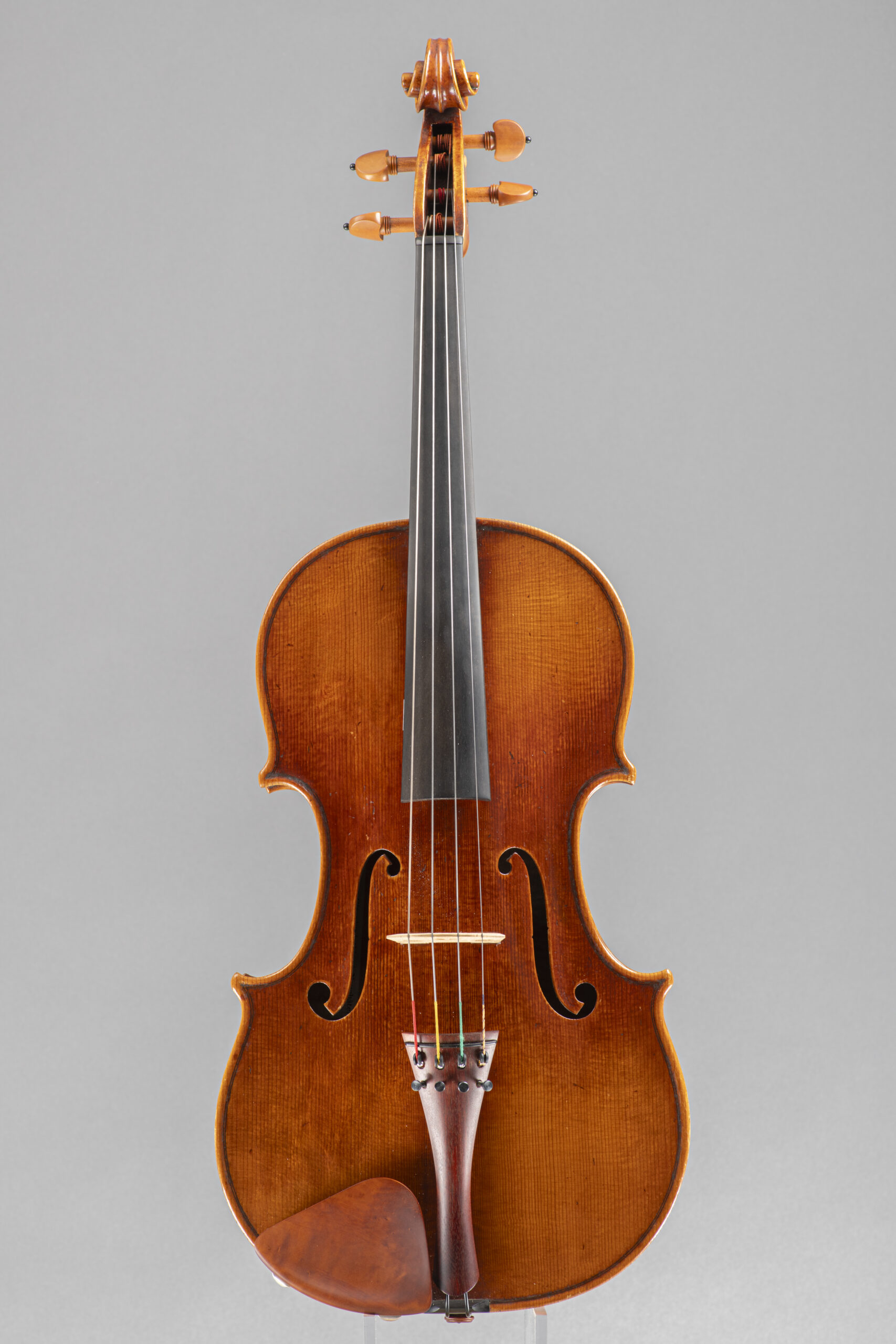 A frontal studio portrait of a wooden violin with a warm-toned, aged finish, featuring an ebony fingerboard and a matching wooden tailpiece and chin rest, centered against a plain grey background.