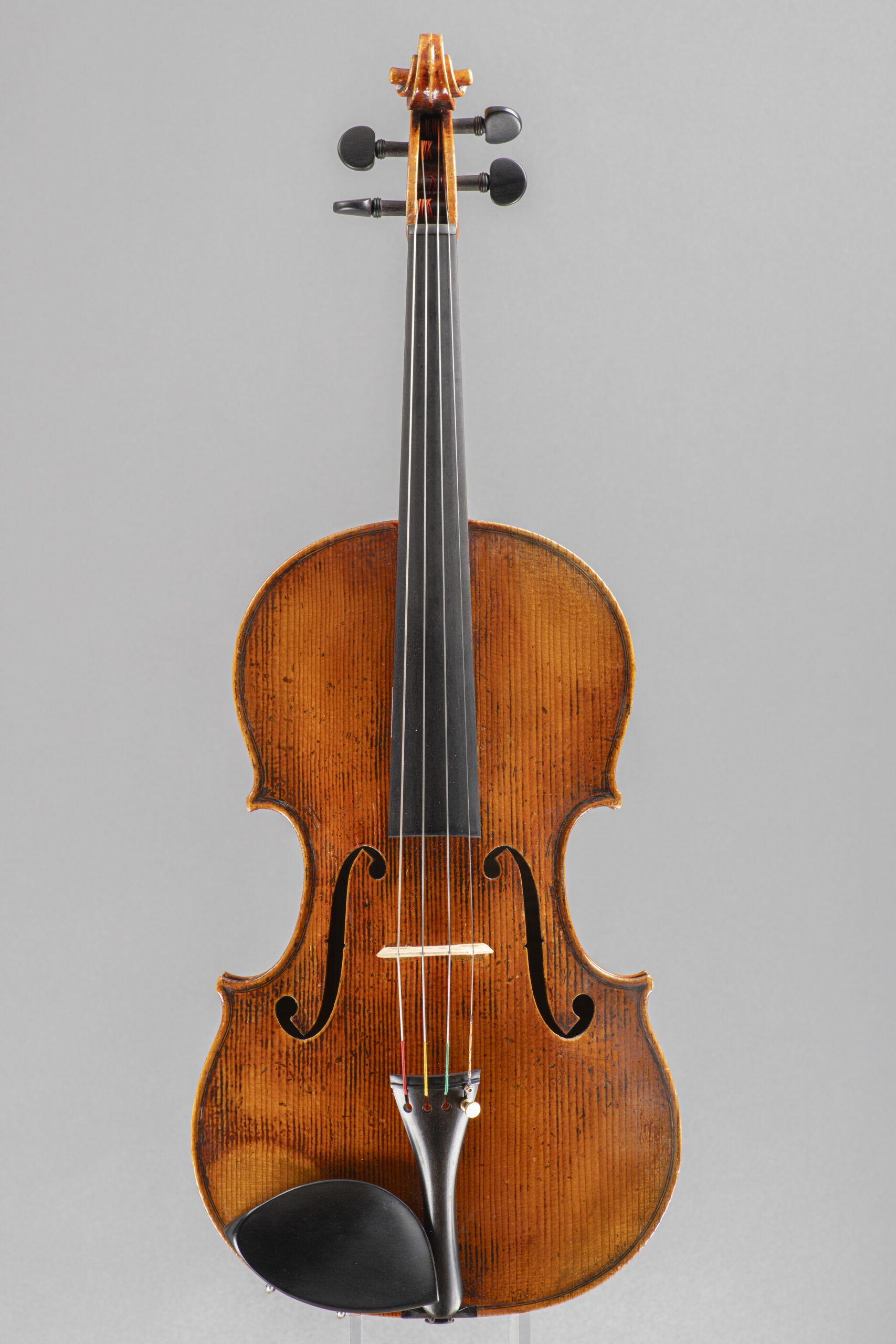 An upright, studio-lit portrait of an aged, wooden violin, featuring a textured brown varnish, a black ebony fingerboard, and a matching black chin rest, centered against a plain grey background.